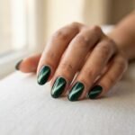 Close-up of a finished cat eye nail manicure on a medium skin tone hand showing a deep emerald green magnetic gel stripe under soft natural window light.