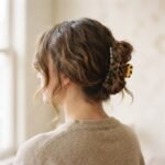 A close-up of a woman with medium brown wavy hair wearing a casually pinned butterfly bun claw clip hairstyle with face-framing wisps, shot in soft natural window light.
