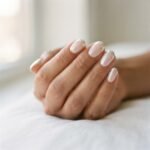 Close-up of a chrome glazed-donut French tip manicure on medium-toned hands with a mirror-finish white pearl tip and sheer nude base, photographed in soft natural window light.