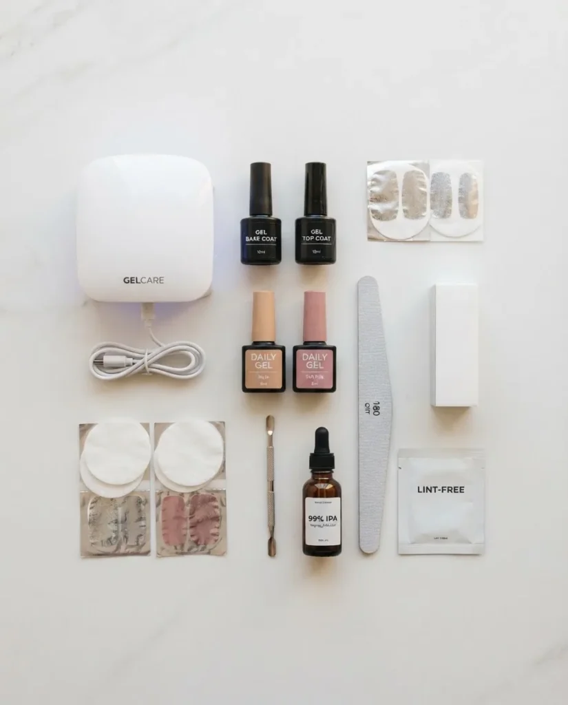 Flat lay of at-home gel manicure beginner tools including an LED lamp, base coat, gel polish bottles, a nail file, buffer block, cuticle pusher, and lint-free wipes on a white surface