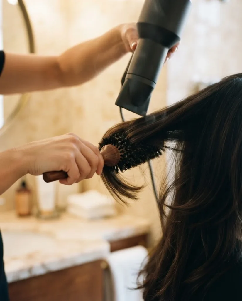  Close-up of hands using a round brush and blow dryer to style long layered hair section by section with warm bathroom vanity lighting.