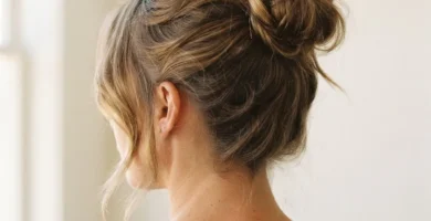 Close-up of a woman's high messy bun hairstyle with soft face-framing pieces, shot in warm natural window light against a neutral background.
