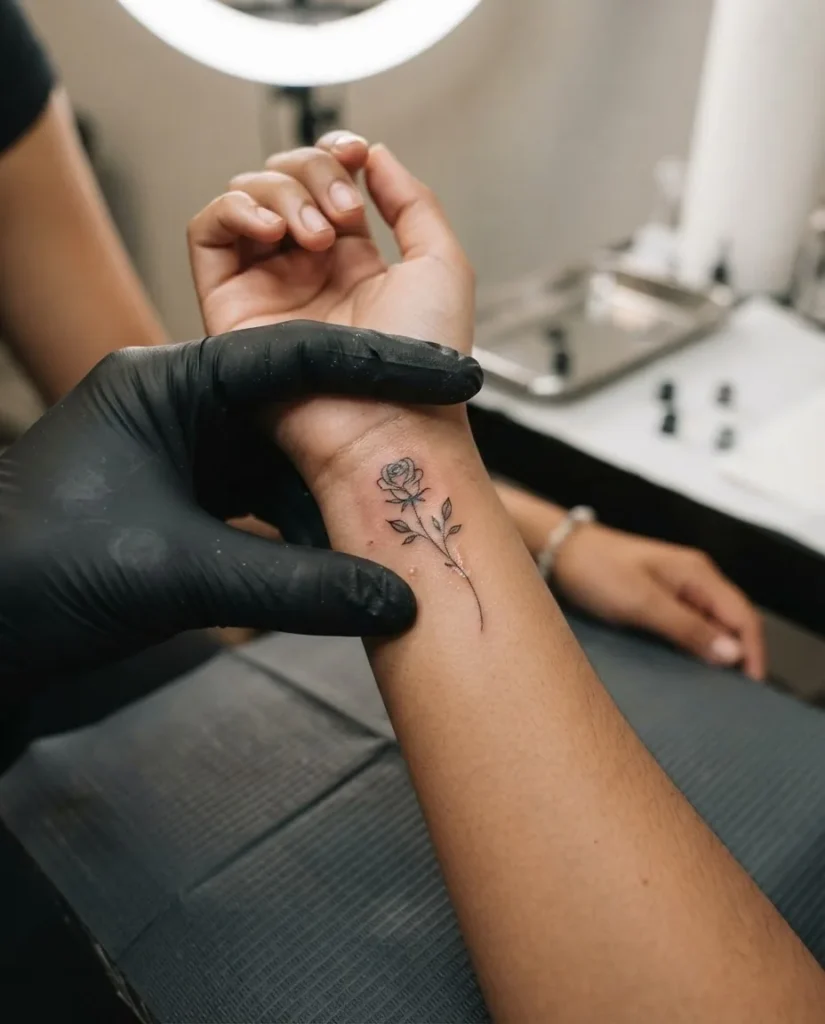 Extreme close-up of a fresh fine line rose stem tattoo on the inner wrist of a woman with medium brown skin tone, needle detail visible on clean skin under soft warm studio lighting.